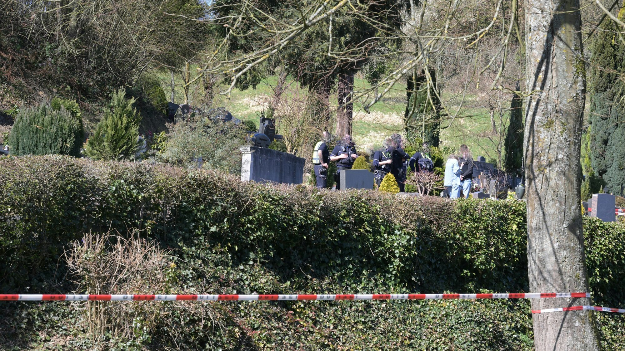 Ein Friedhof ist mit Polizei-Flatterband abgesperrt, zwischen Gräbern stehen unter anderem Polizeikräfte.
