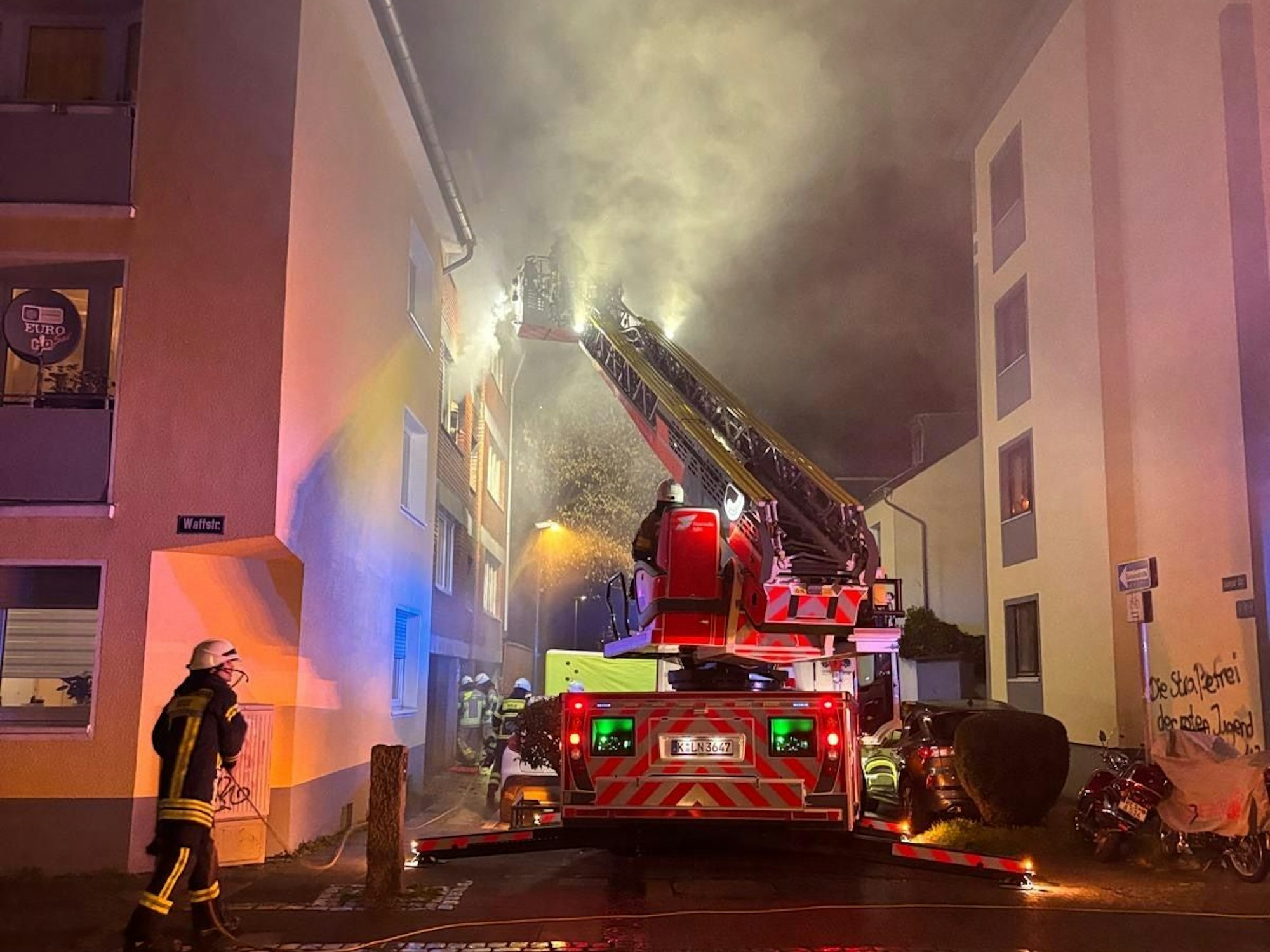 Aus einem Fenster im zweiten Stock dringt starker Rauch, vor dem Haus steht ein Fahrzeug der Feuerwehr, sind mehrere Wehrleute im Einsatz.