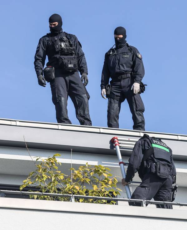 Zwei vermummte Polizisten stehen auf einem Hausdach, ein dritter klettert eine Leiter runter auf einen Balkon.