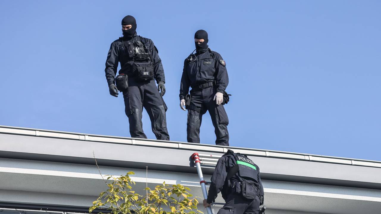Zwei vermummte Polizisten stehen auf einem Hausdach, ein dritter klettert eine Leiter runter auf einen Balkon.