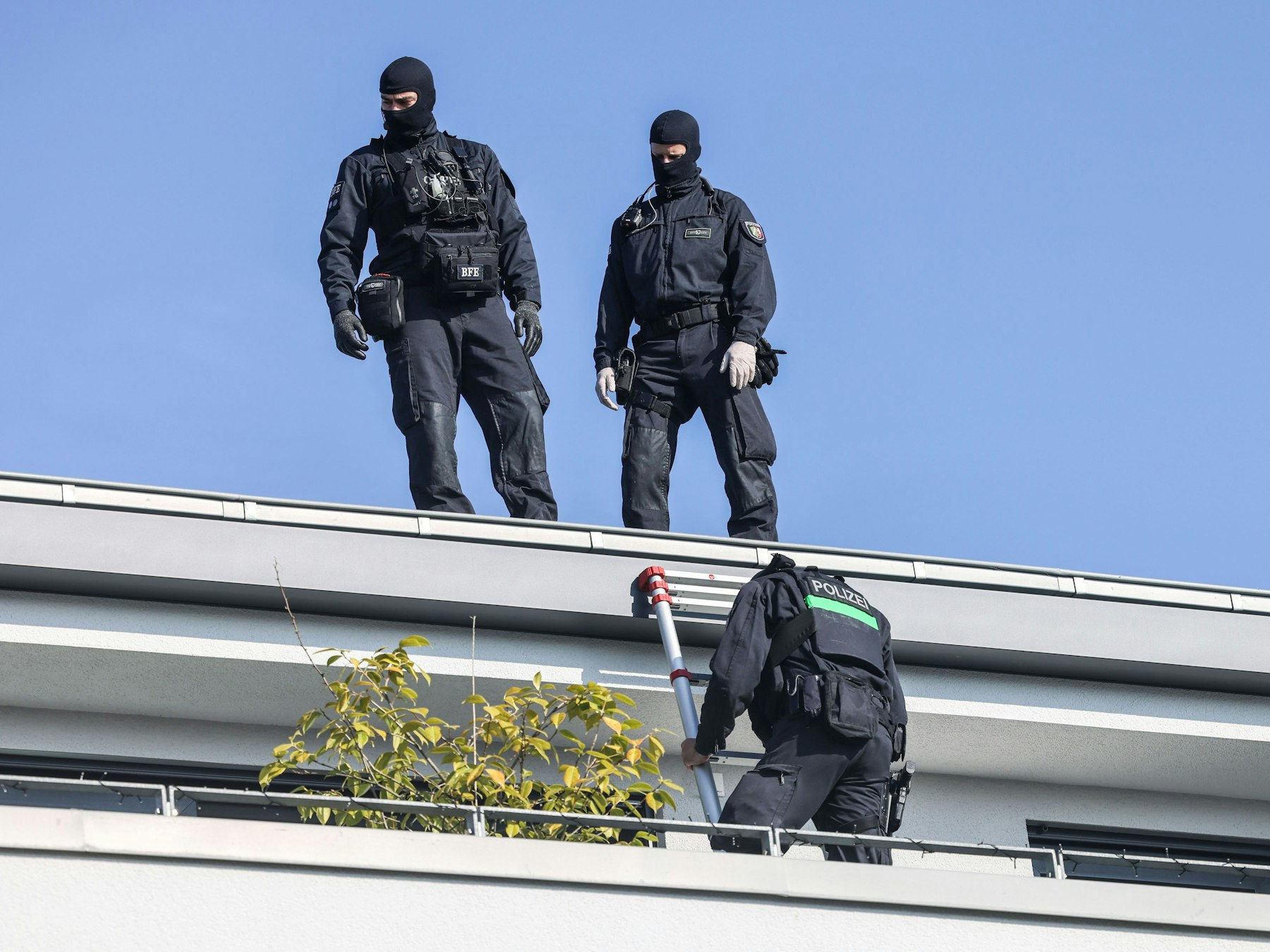 Zwei vermummte Polizisten stehen auf einem Hausdach, ein dritter klettert eine Leiter runter auf einen Balkon.
