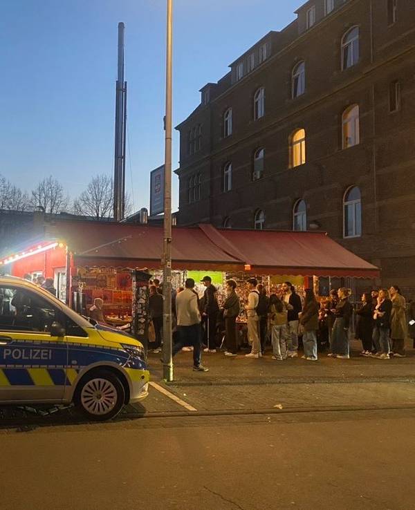 Ein Polizeifahrzeug hält vor einem Imbiss, vor dem die Leute Schlange stehen.
