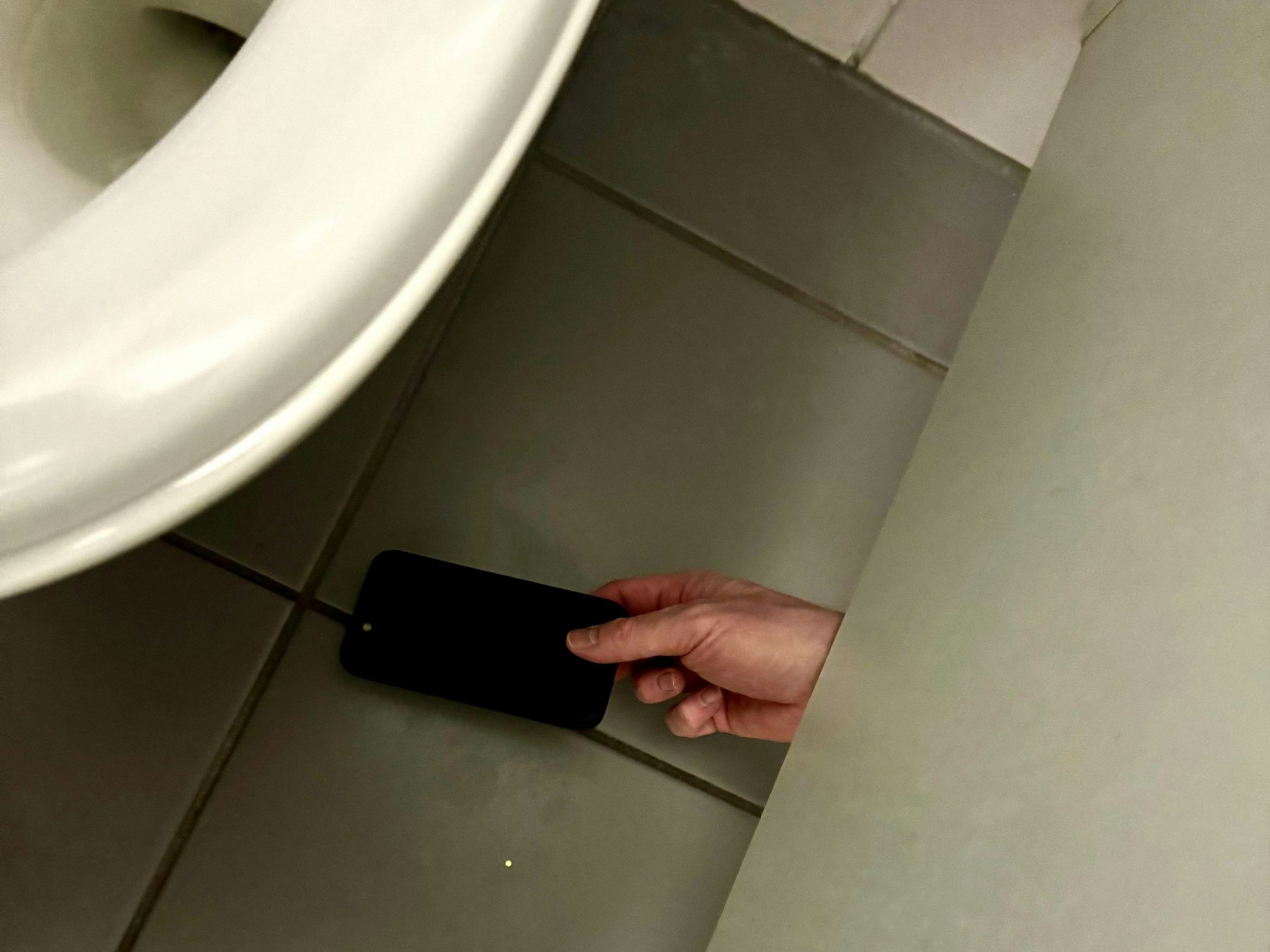 Eine Person hält ein Handy unter der Trennwand einer Toilette durch.