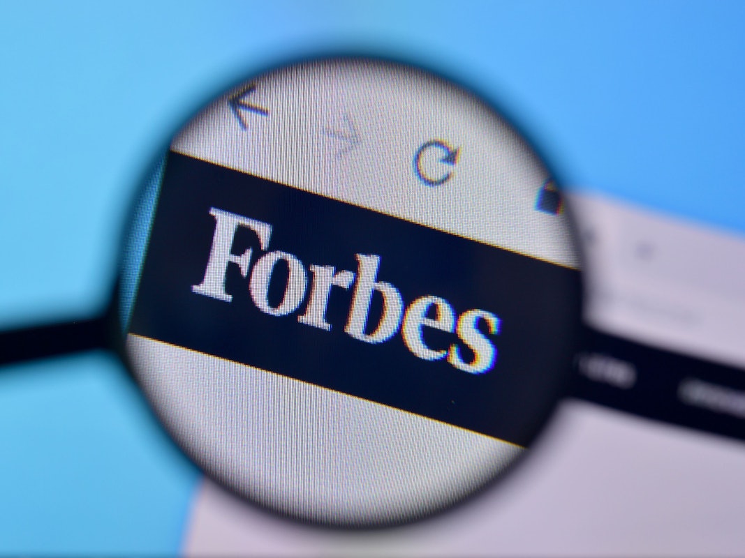 Zu sehen ist eine Lupe die das Logo der „Forbes“-Onlineseite vergrößert.
