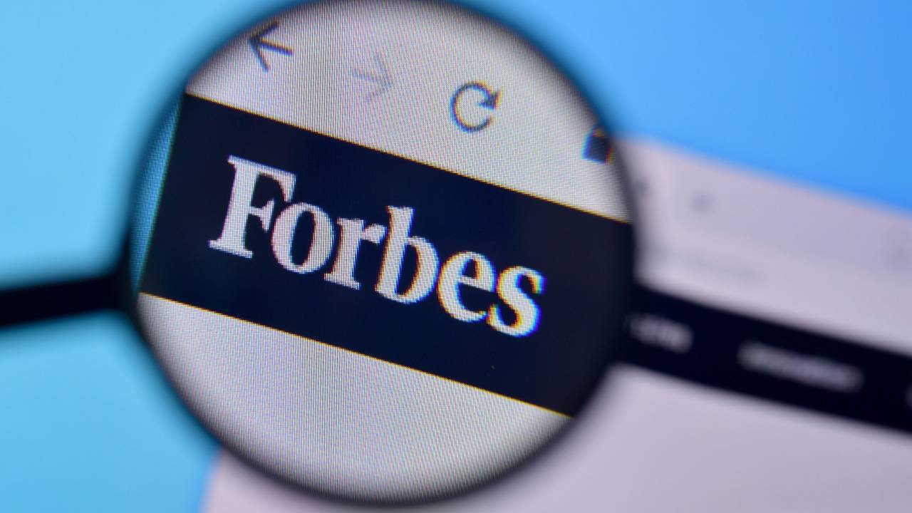 Zu sehen ist eine Lupe die das Logo der „Forbes“-Onlineseite vergrößert.