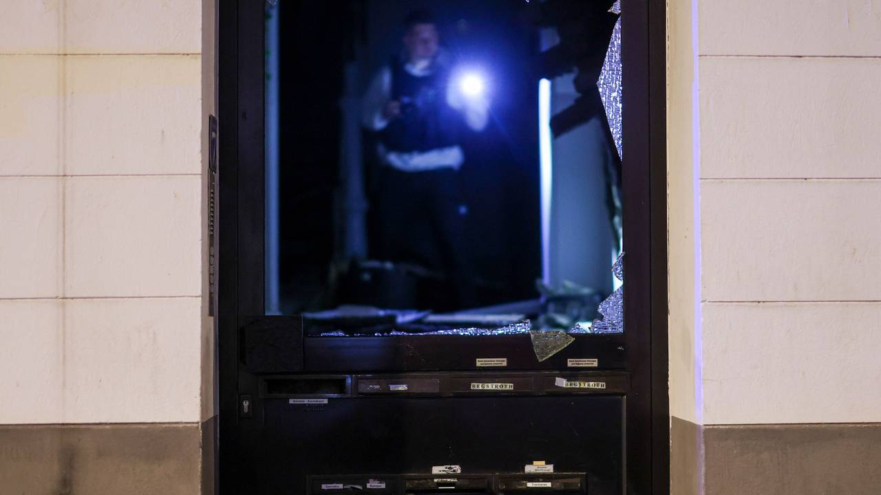 Ein Polizeibeamter der Spurensicherung untersucht im Hausflur den Tatort, durch die Explosion im Erdgeschoss des Hauses wurde die Hauseingangstüre beschädigt.
