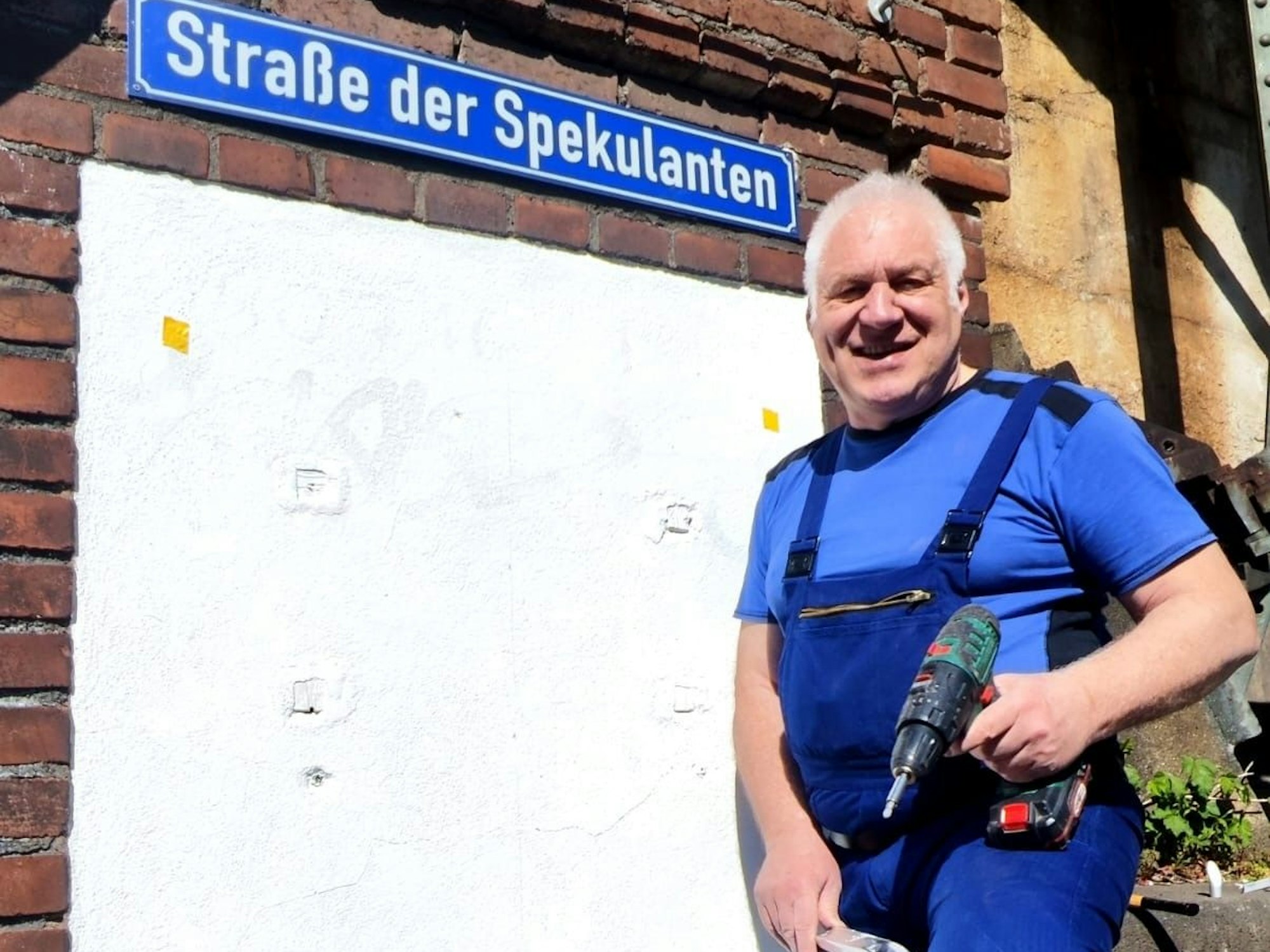 Ein Mann steht mit Bohrmaschine in der Hand vor einem Straßenschild mit der Aufschrift Straße der Spekulanten.