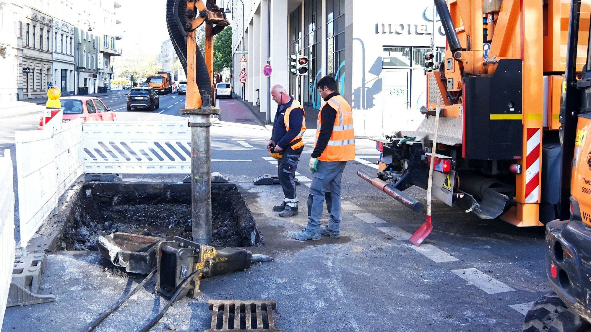 Zwei Männer gucken in eine abgesackte Stelle auf einer Fahrbahn, hinter ihnen steht ein Bagger.