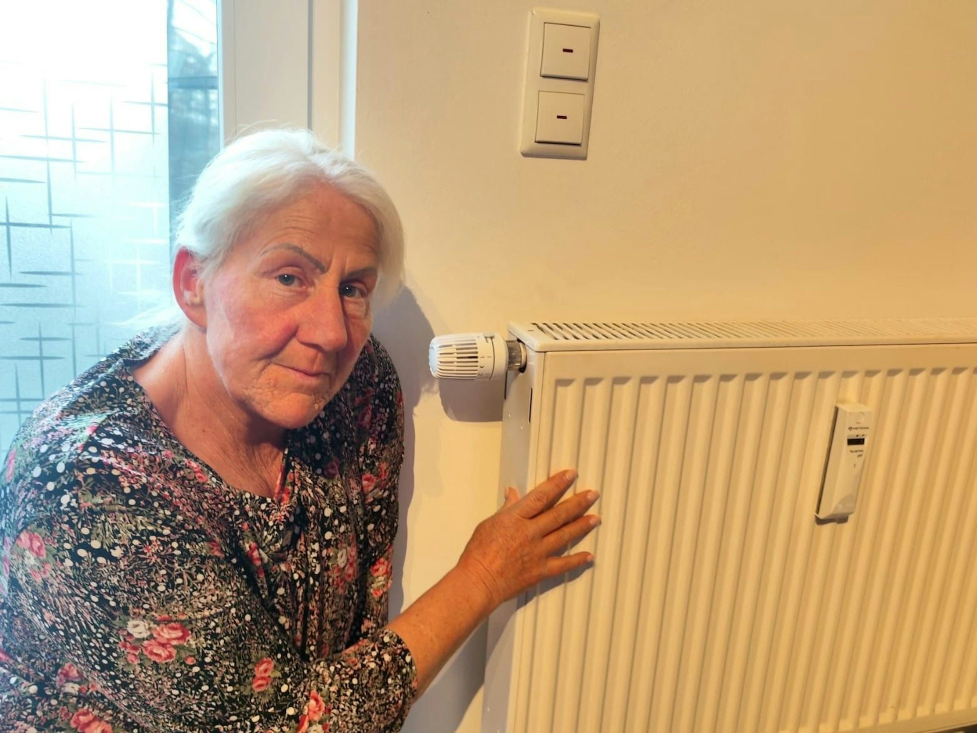 Eine ältere Frau hockt neben einem Heizkörper in ihrer Wohnung.