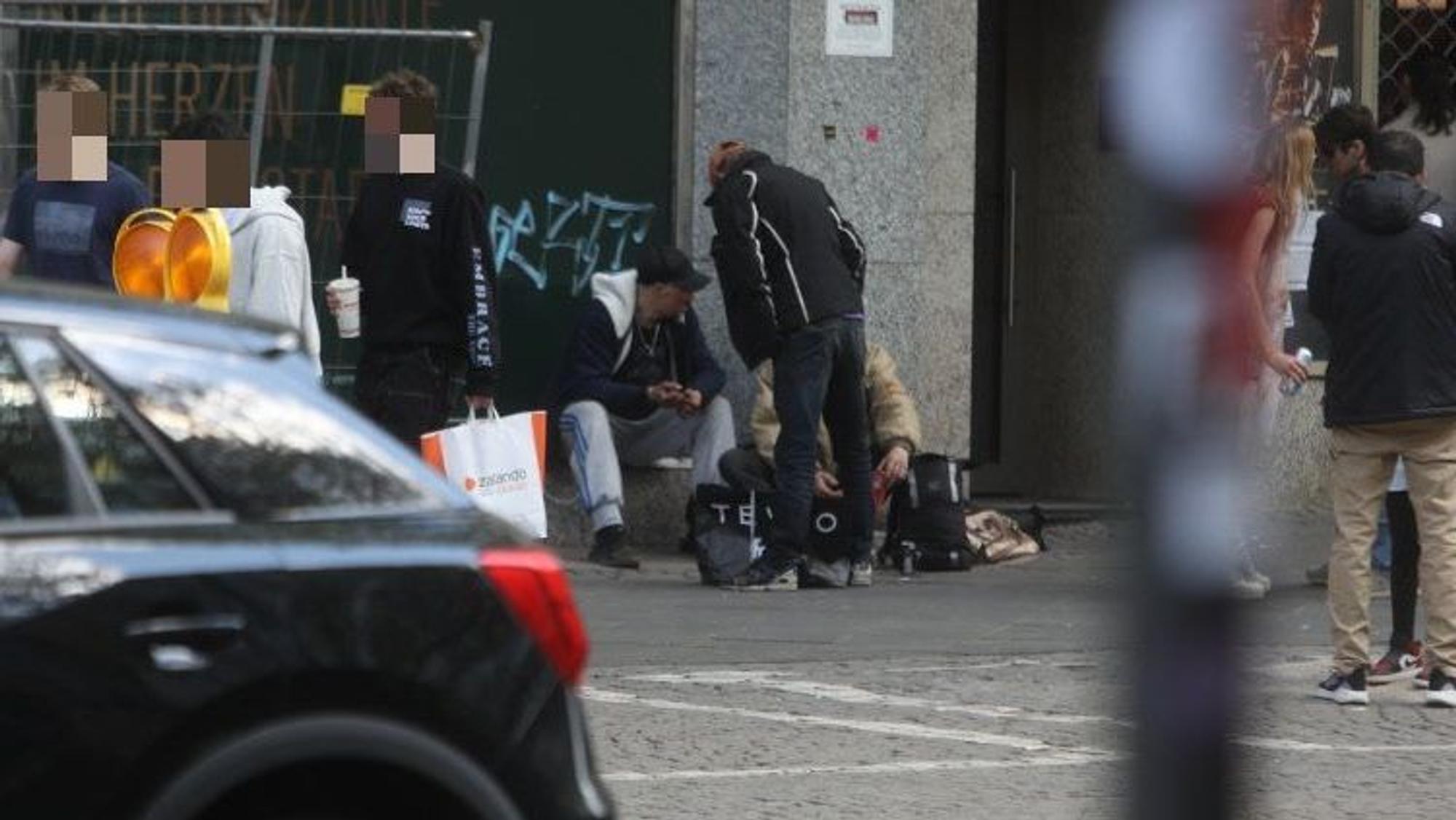 Drei Männer hängen vor einem Gebäude ab, junge Passanten gehen an ihnen vorbei.
