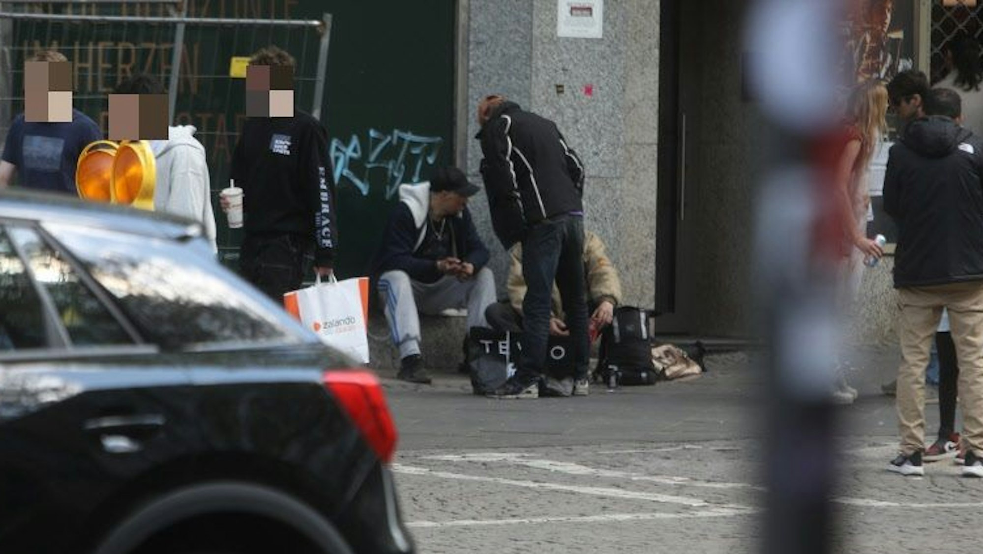 Drei Männer hängen vor einem Gebäude ab, junge Passanten gehen an ihnen vorbei.