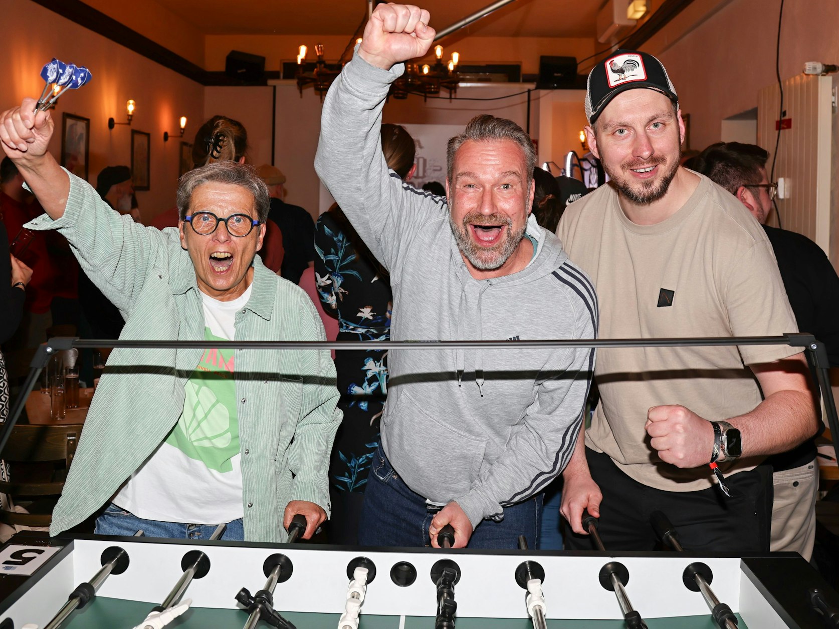 Zwei Männer und eine Frau stehen am Kickertisch und jubeln.