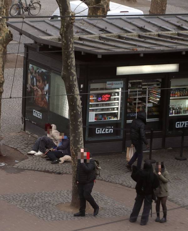 Menschen laufen über einen Platz, an einem Kiosk sitzen zwei Personen auf dem Boden.