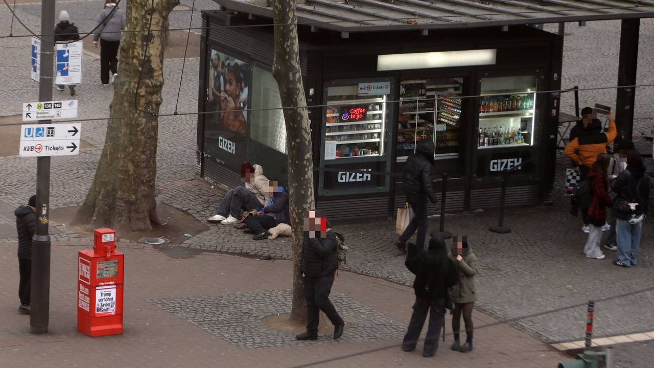 Menschen laufen über einen Platz, an einem Kiosk sitzen zwei Personen auf dem Boden.