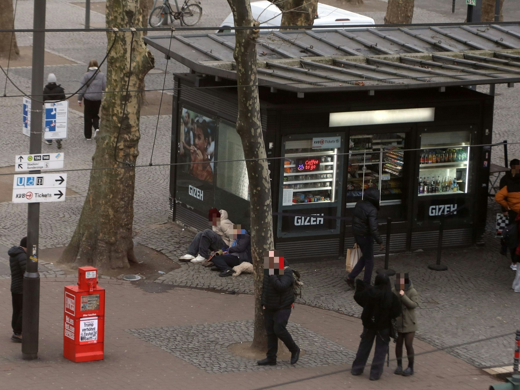 Menschen laufen über einen Platz, an einem Kiosk sitzen zwei Personen auf dem Boden.