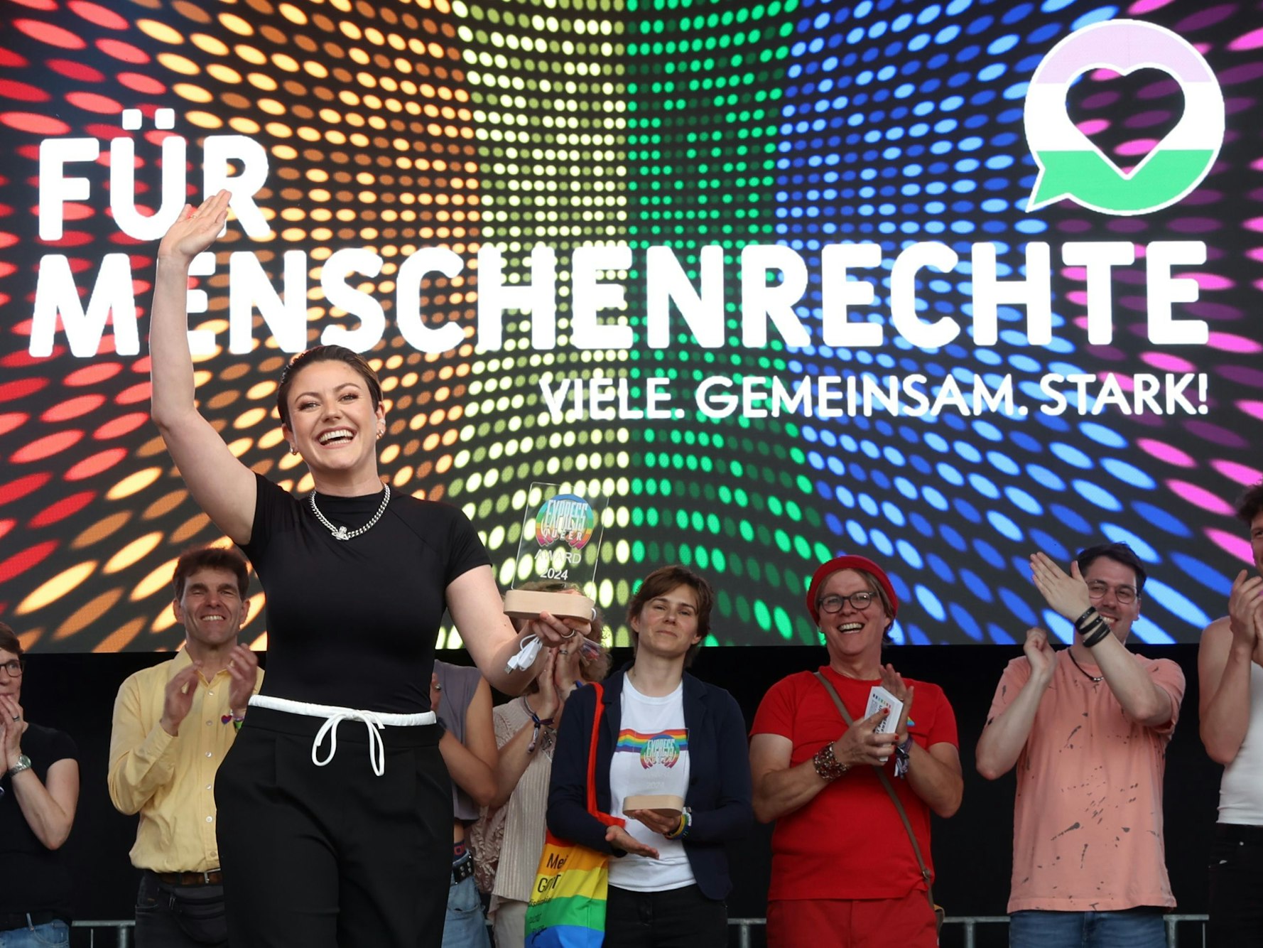 Eine Frau hält einen Preis in der Hand, mit der anderen Hand winkt sie. Hinter ihr applaudieren mehrere Menschen.