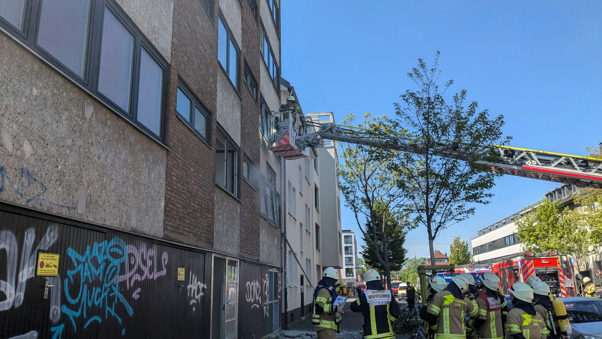 Feuerwehrleute stehen vor einem Mehrfamilienhaus und blicken hoch zum ersten Stock. Dorthin sind Kräfte auf der Drehleiter im Einsatz.