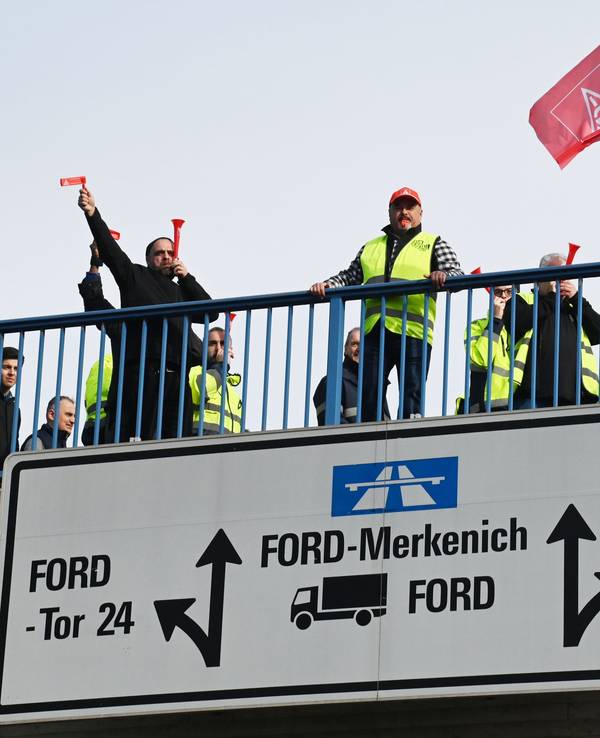 Mehrere Personen stehen auf einer Brücke zum Fordwerk, ein Mann schwenkt eine IG-Metall-Fahne.