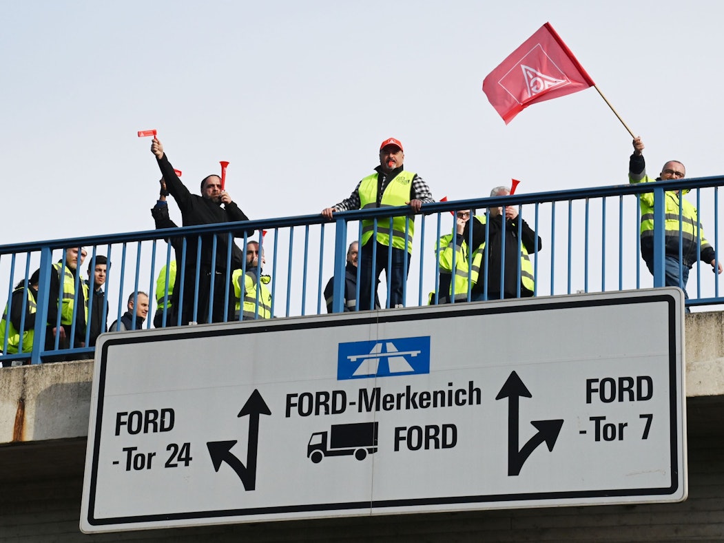 Mehrere Personen stehen auf einer Brücke zum Fordwerk, ein Mann schwenkt eine IG-Metall-Fahne.