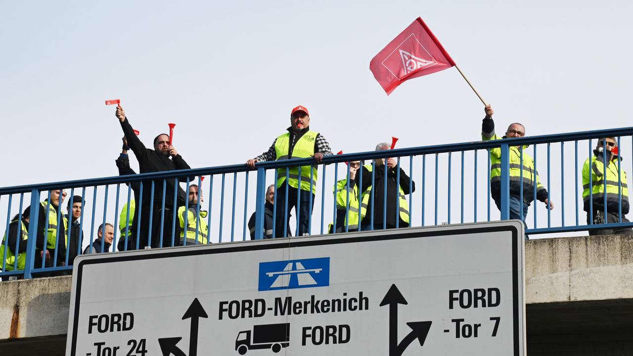 Mehrere Personen stehen auf einer Brücke zum Fordwerk, ein Mann schwenkt eine IG-Metall-Fahne.