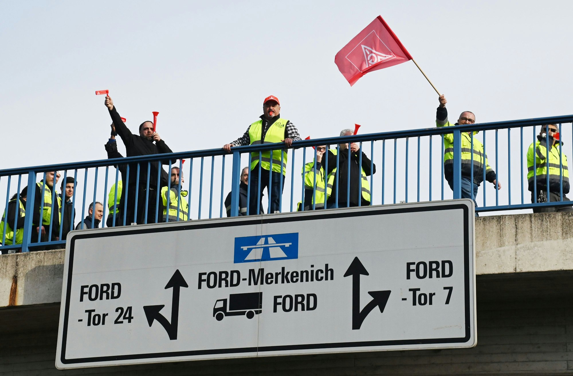 Mehrere Personen stehen auf einer Brücke zum Fordwerk, ein Mann schwenkt eine IG-Metall-Fahne.