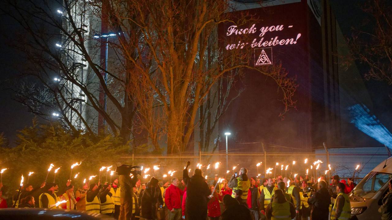 Ford-Mitarbeiter stehen mit Fackeln auf dem Gehweg vor einem Gebäude der Fordwerke, auf dem die Worte „Fuck you – wir bleiben" projiziert wird.