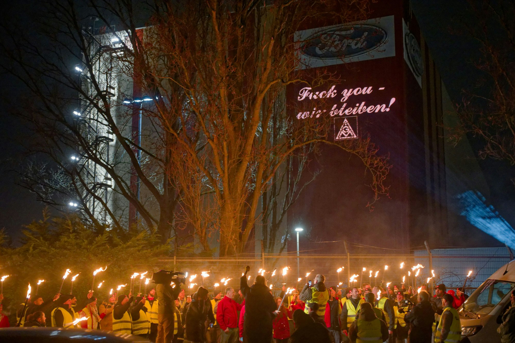 Ford-Mitarbeiter stehen mit Fackeln auf dem Gehweg vor einem Gebäude der Fordwerke, auf dem die Worte „Fuck you – wir bleiben" projiziert wird.
