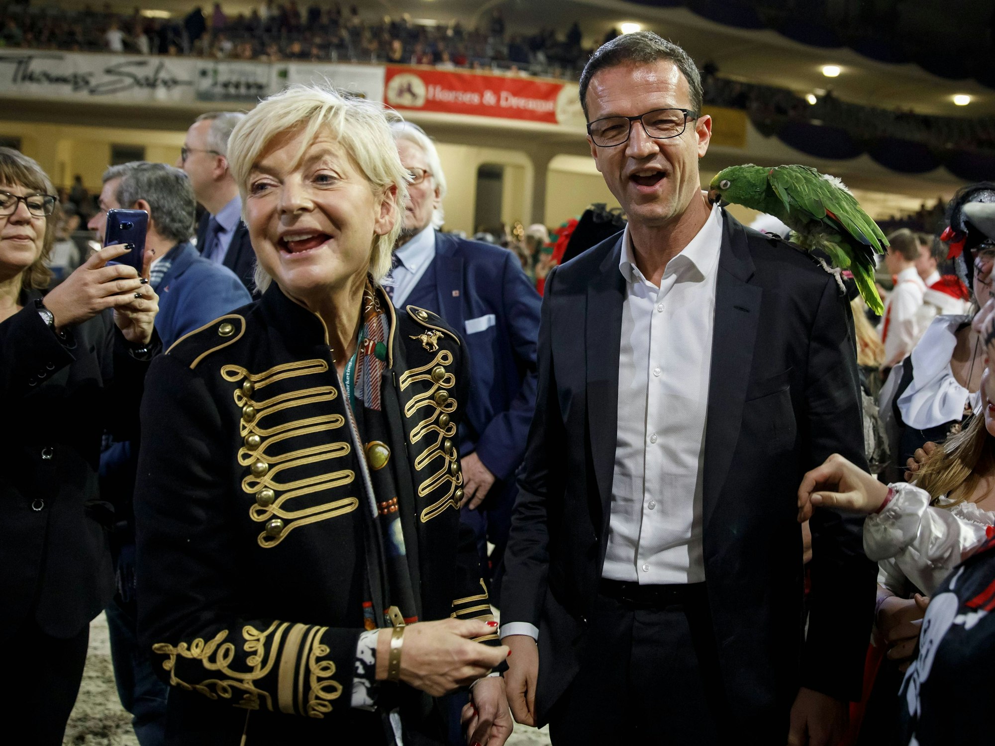 Ann Kathrin Linsenhoff steht neben Fred Bobic, dem ein grüner Papagei in den Hemdkragen beißt.