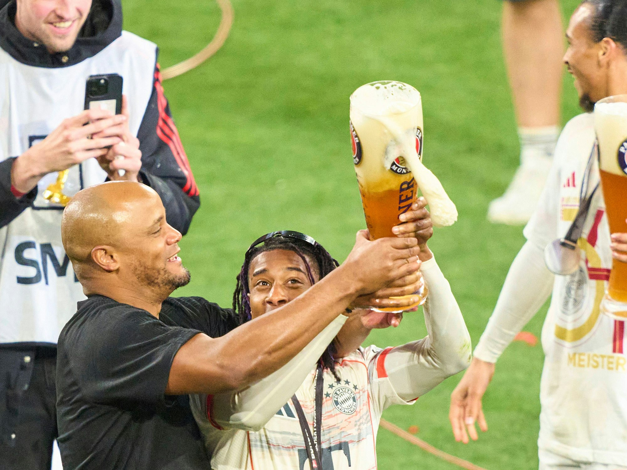Vincent Kompany und Michael Olise streiten sich spielerisch um ein übergroßes Weizenbier.