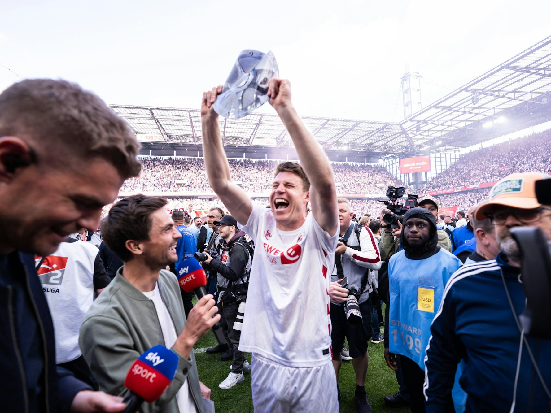 Dominique Heintz hält eine Meisterschale aus Papier hoch. Max Zielke und Simon Terodde von Pay-TV-Sender Sky stehen daneben.