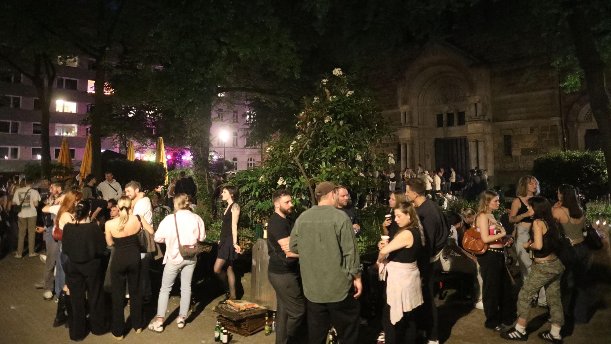 Auf einem Platz stehen in der Nacht junge Menschen in Gruppen, trinken Bier und unterhalten sich.
