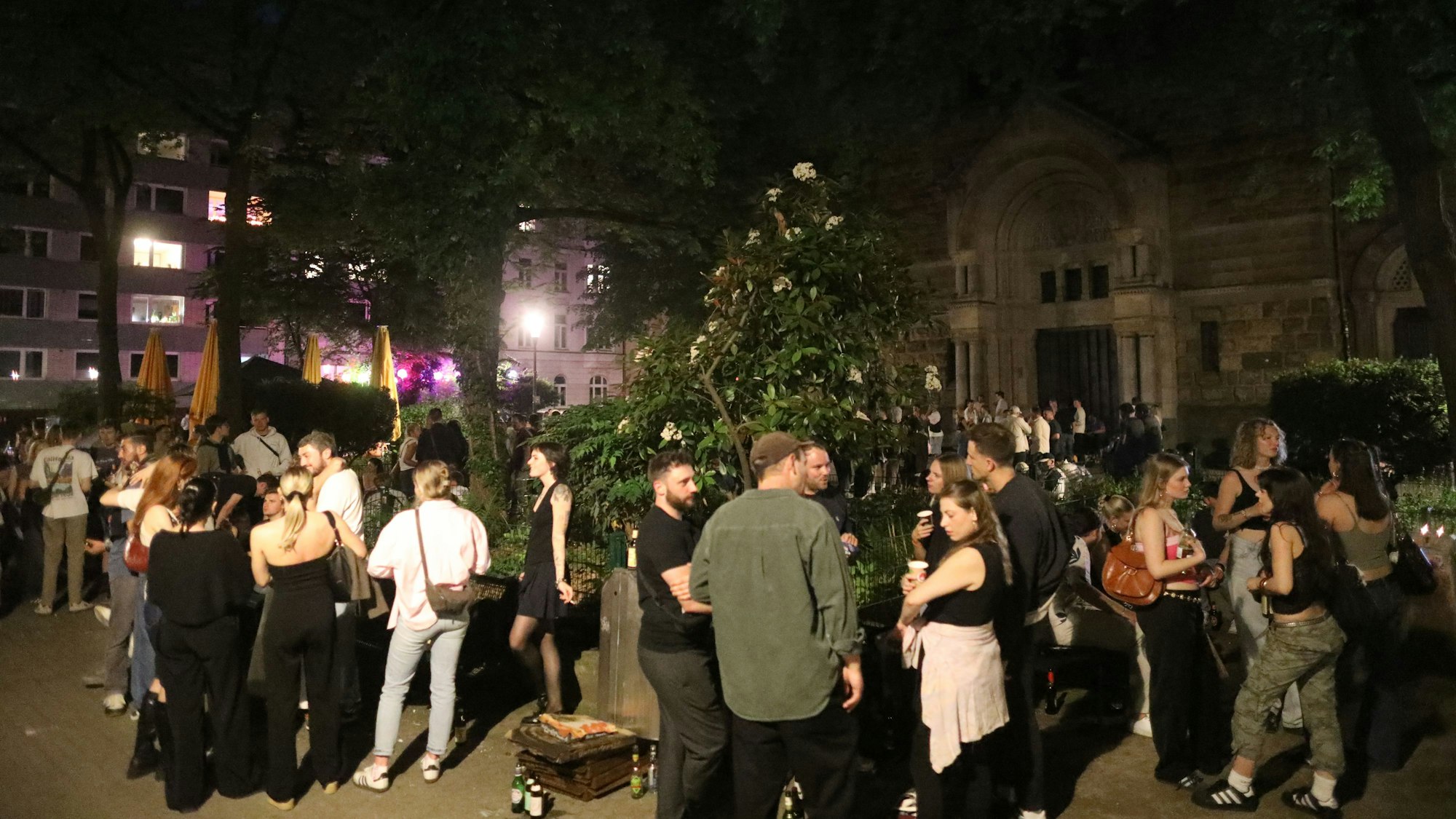 Auf einem Platz stehen in der Nacht junge Menschen in Gruppen, trinken Bier und unterhalten sich.