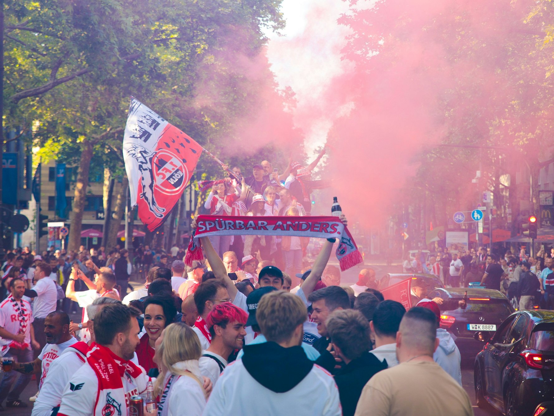 Menschen mit Fanschals des 1. FC Köln knubbeln sich auf einer Straße.