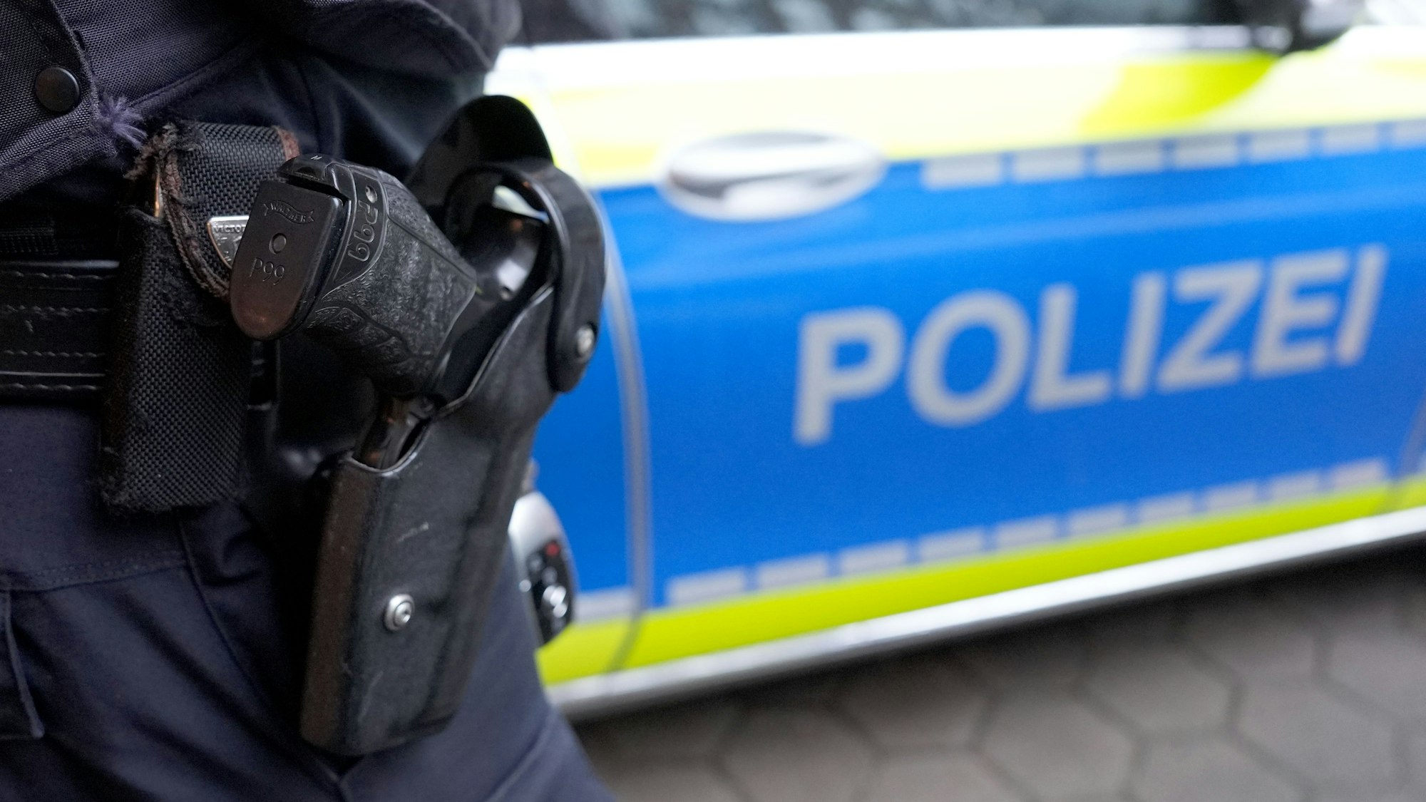Vor einem Streifenwagen steht ein Polizist, von dem man nur die Waffe sieht.