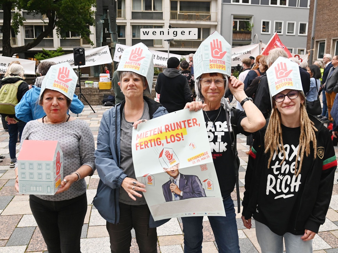 Vier Frauen tragen auf dem Kopf Papphüte mit der Aufschrift Mietenstopp, zwei halten zudem ein Plakat in Händen mit der Aufschrift „Mieten runter, fertig, los!“