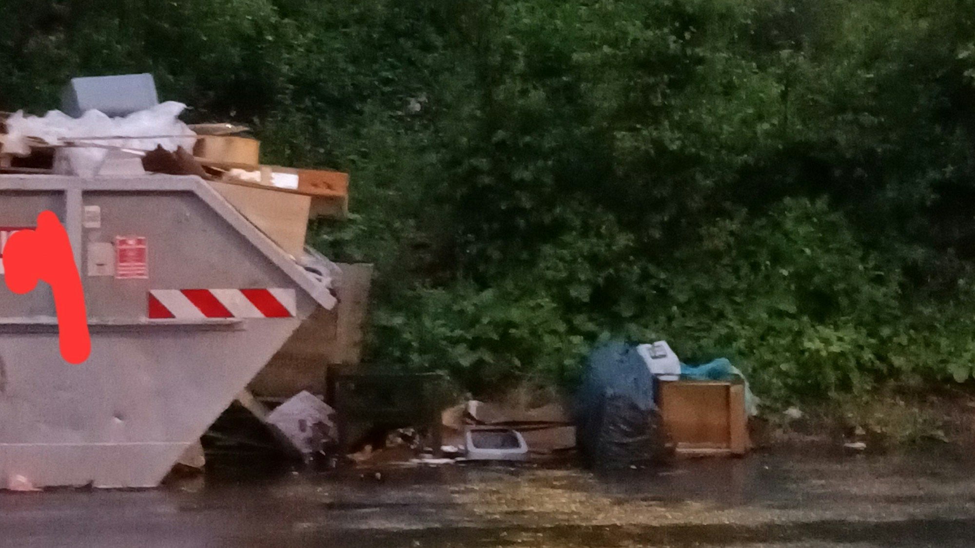 An einer Straße steht ein übervoller Müllcontainer, auf dem Boden liegt ebenfalls Müll.