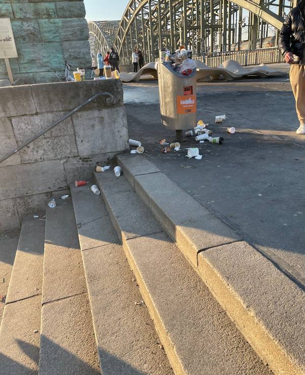 Um einen Mülleimer herum liegen To-Go-Becher auf dem Boden und auf den Stufen einer angrenzenden Treppe.