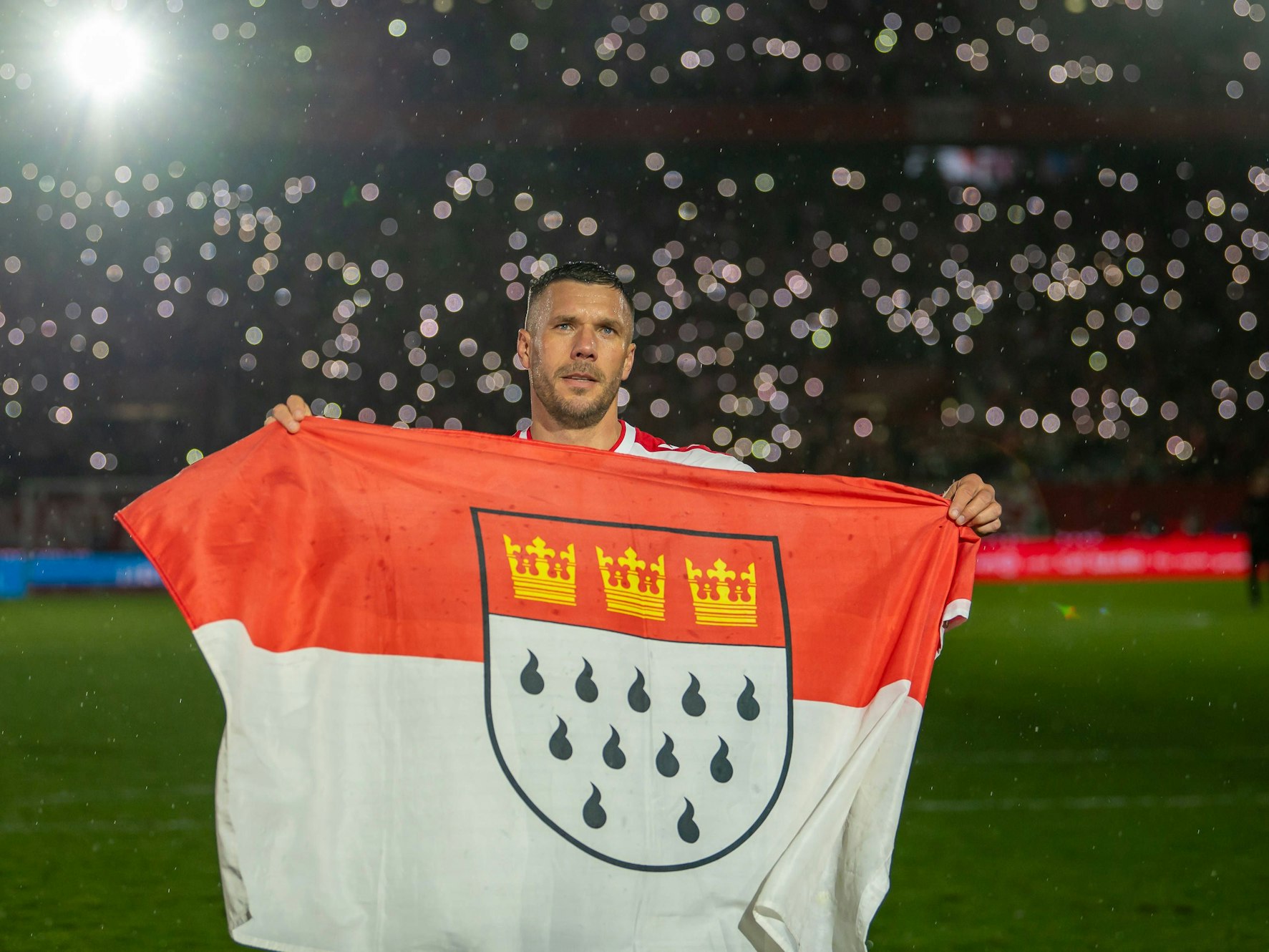 Lukas Podolski steht mit einer großen Köln-Fahne auf dem Platz.