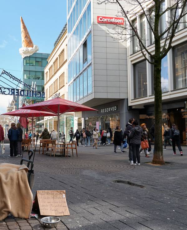 Eine Person sitzt in einer Einkaufsstraße und bittet auf einem Pappschild um Geld.