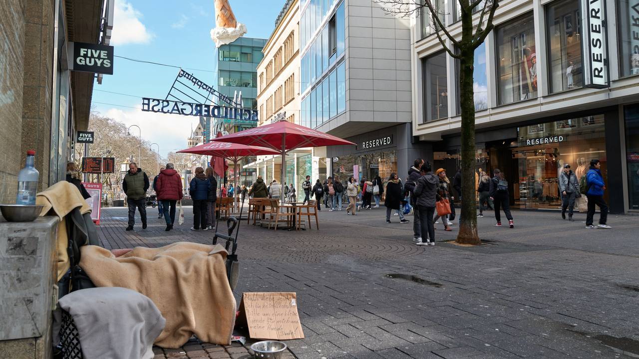 Eine Person sitzt in einer Einkaufsstraße und bittet auf einem Pappschild um Geld.