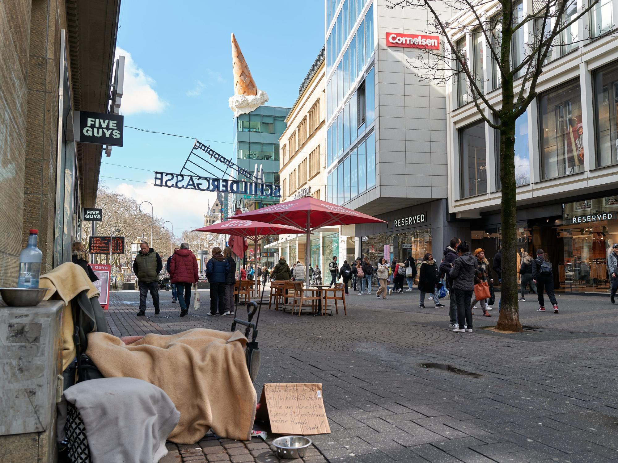 Eine Person sitzt in einer Einkaufsstraße und bittet auf einem Pappschild um Geld.