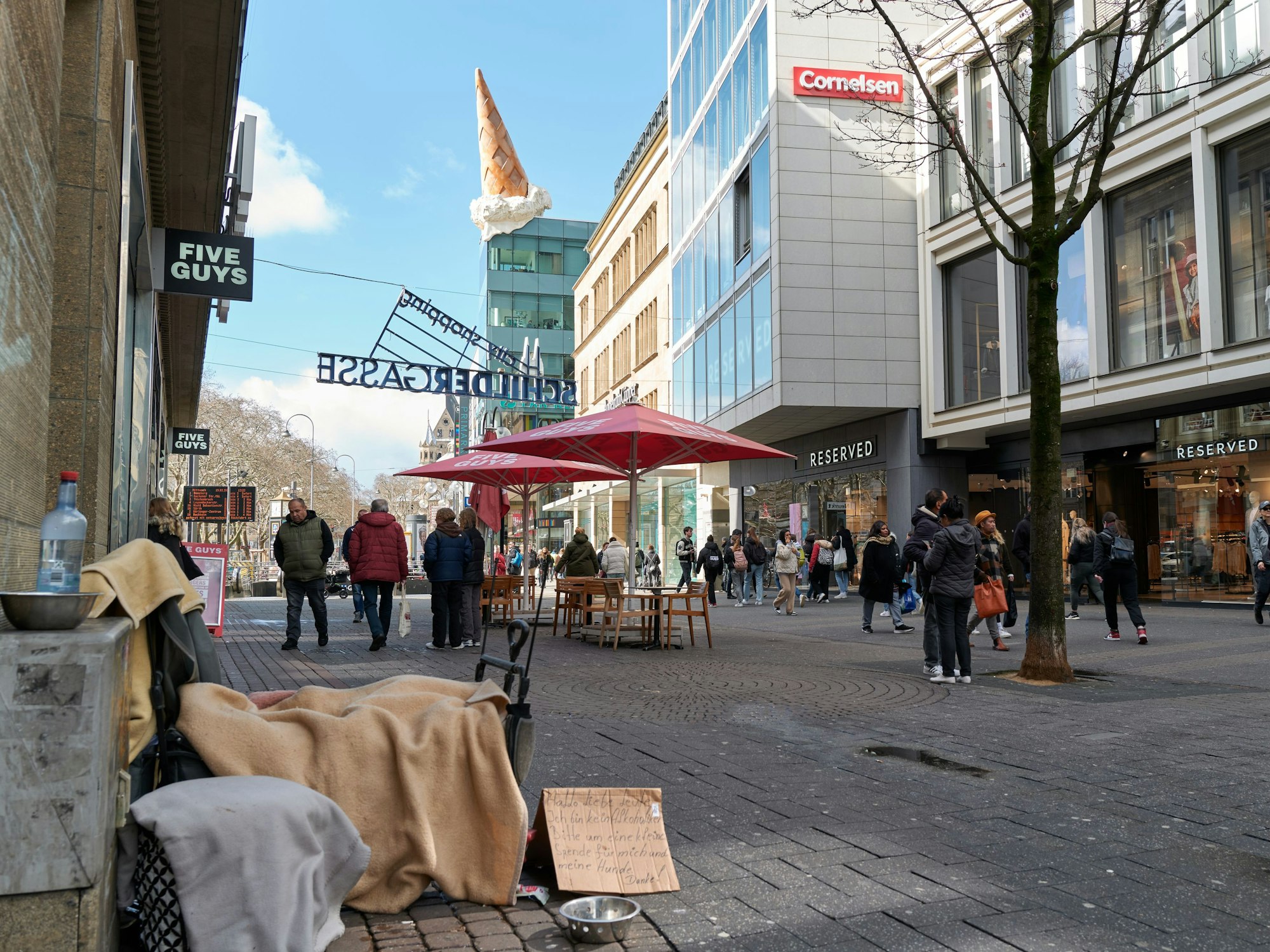 Eine Person sitzt in einer Einkaufsstraße und bittet auf einem Pappschild um Geld.