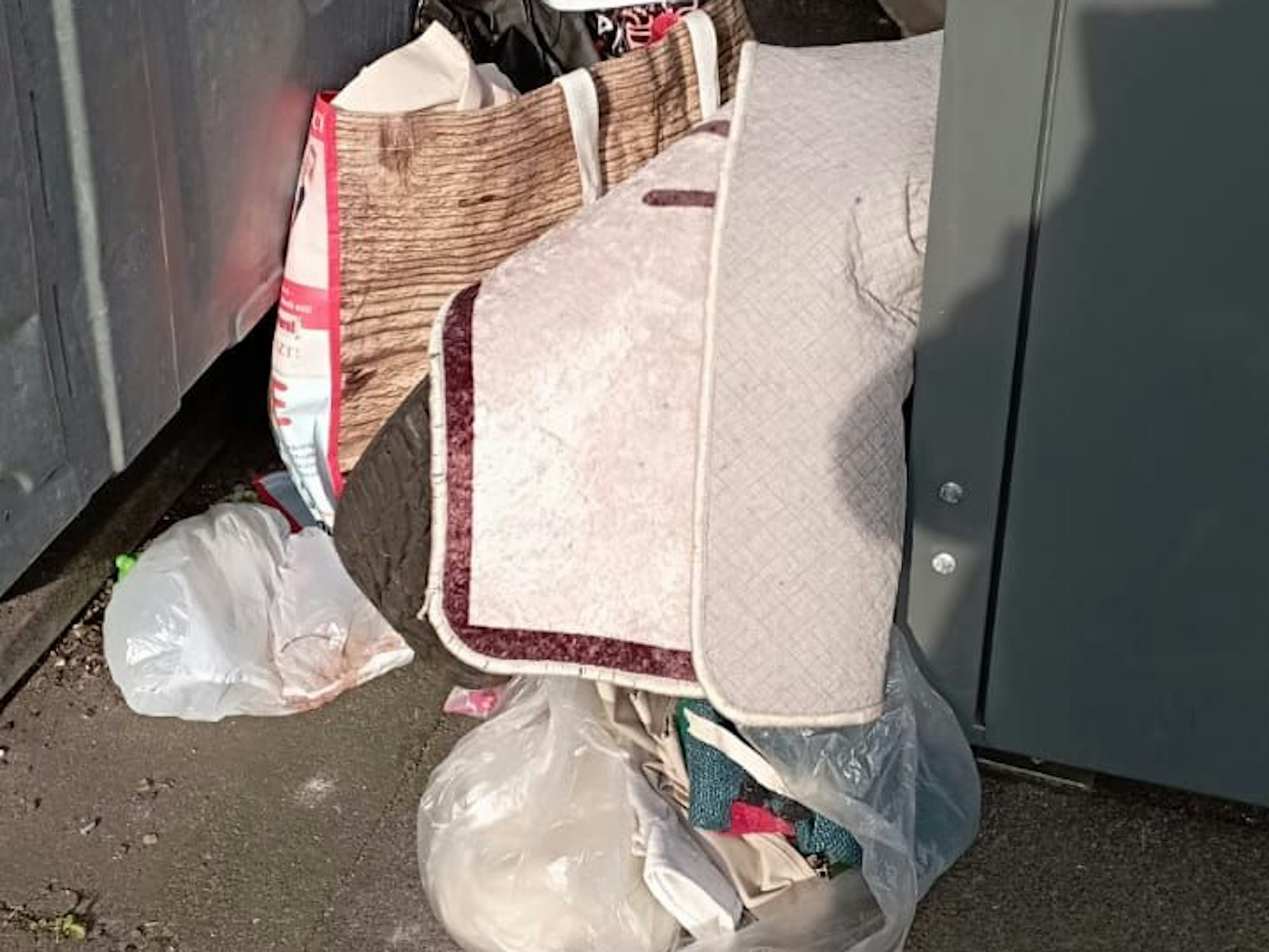 Zwischen zwei Containern stehen gefüllt Plastiktüten, darüber liegt ein Teppich.