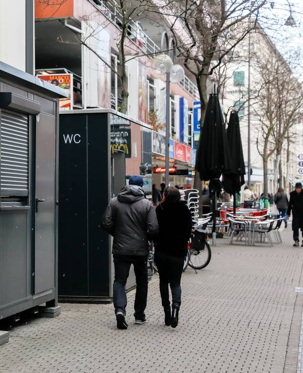 Vor einem Toilettenhäuschen mit WC-Aufschrift steht ein weiterer Container, dieser hat jedoch ein großes Fenster.