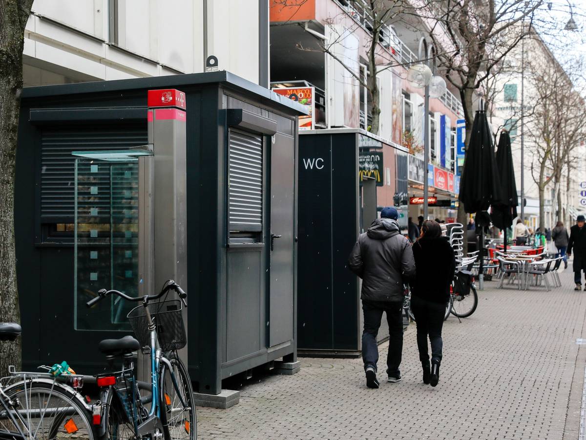 Vor einem Toilettenhäuschen mit WC-Aufschrift steht ein weiterer Container, dieser hat jedoch ein großes Fenster.