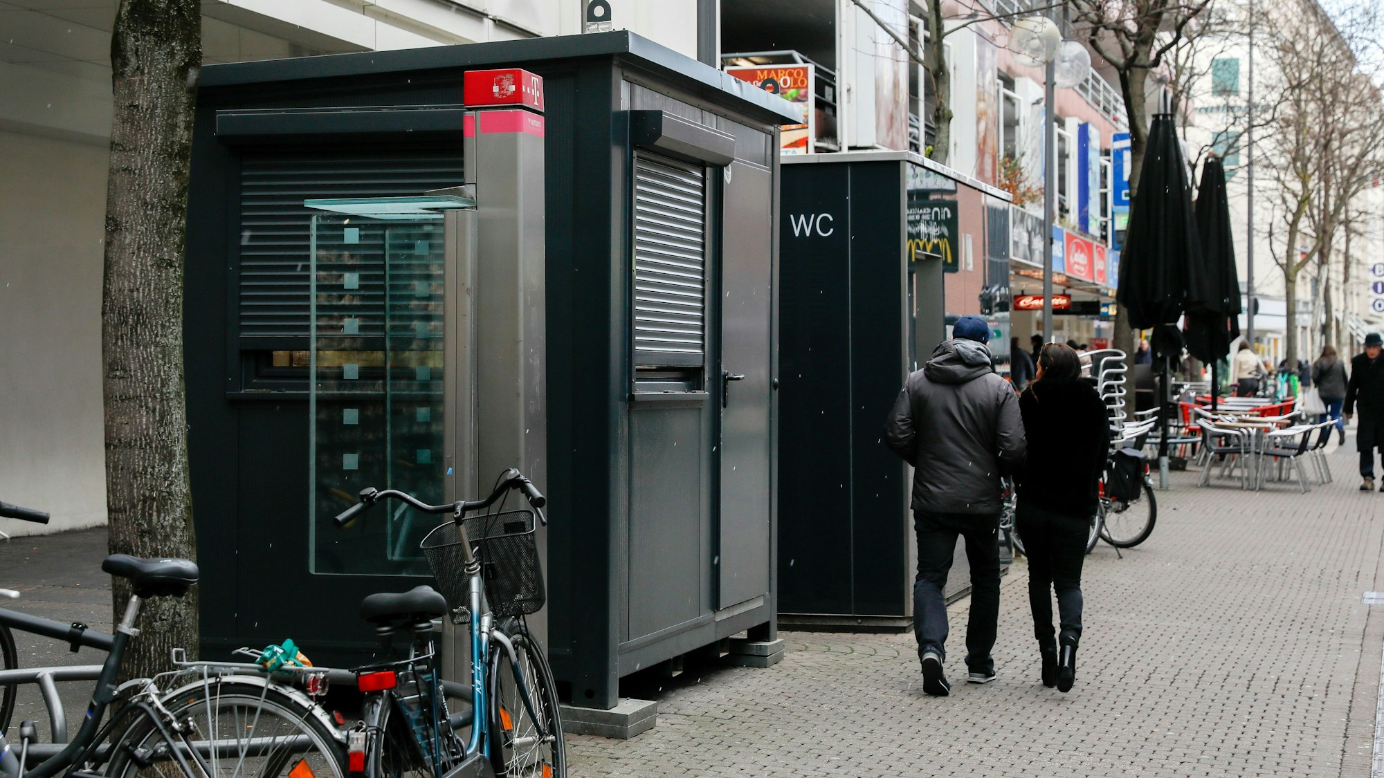 Vor einem Toilettenhäuschen mit WC-Aufschrift steht ein weiterer Container, dieser hat jedoch ein großes Fenster.