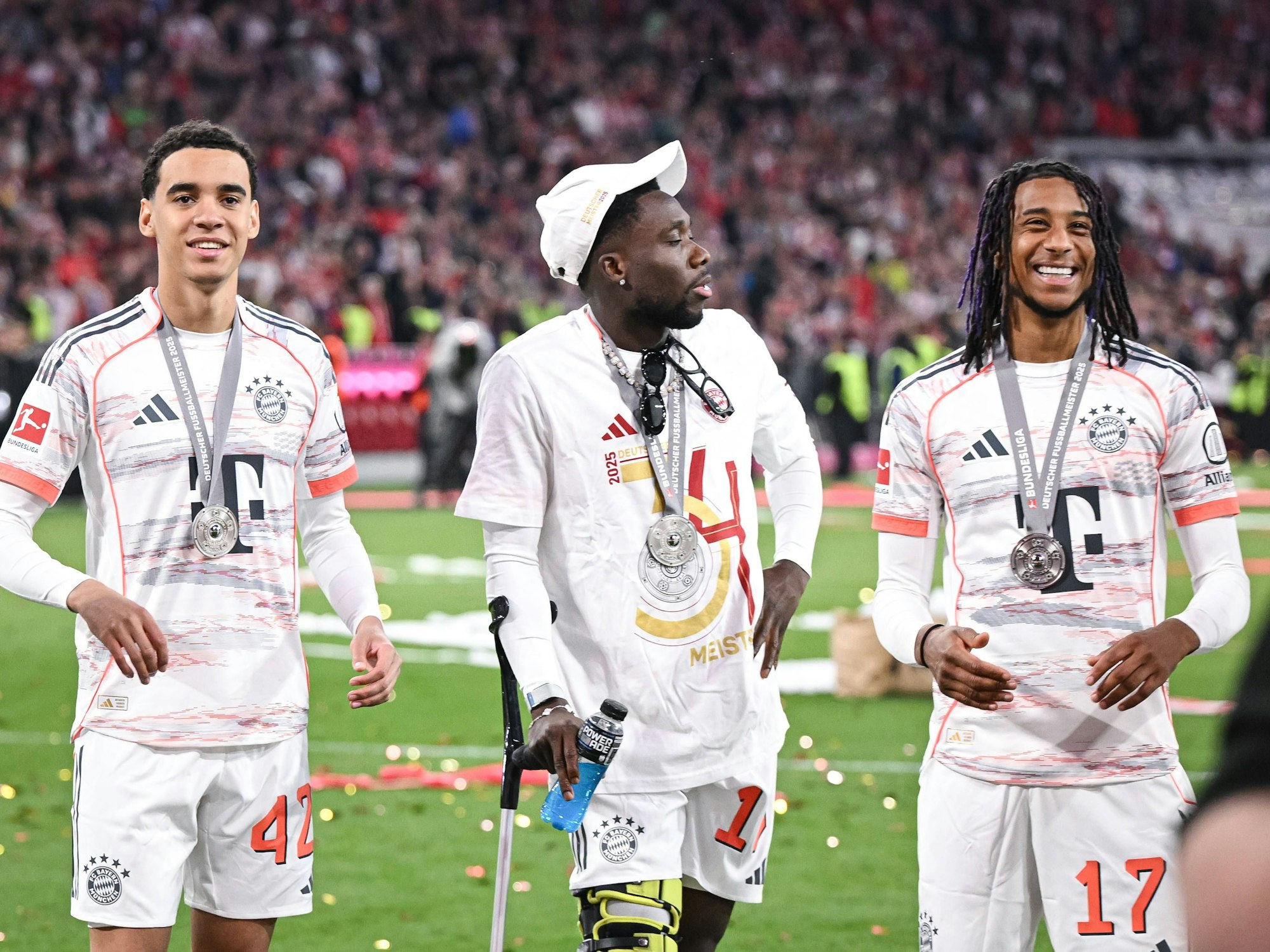 Alphonso Davies steht bei der Schalenübergabe mit einer Krücke zwischen Jamal Musiala und Michael Olise.