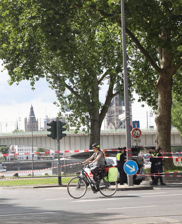 Fahrradfahrende radeln an einem abgesperrten Bereich in Rheinnähe vorbei. Im Hintergrund führt eine Brücke über den Fluss.