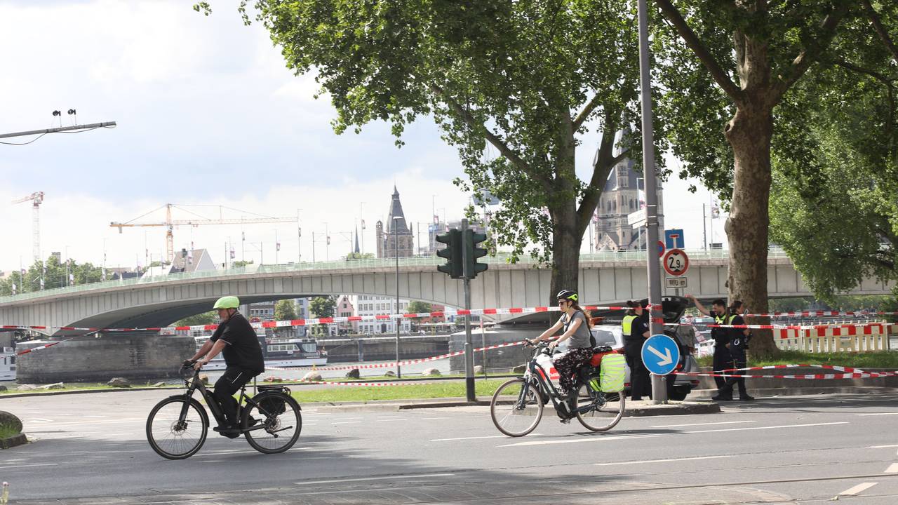 Fahrradfahrende radeln an einem abgesperrten Bereich in Rheinnähe vorbei. Im Hintergrund führt eine Brücke über den Fluss.