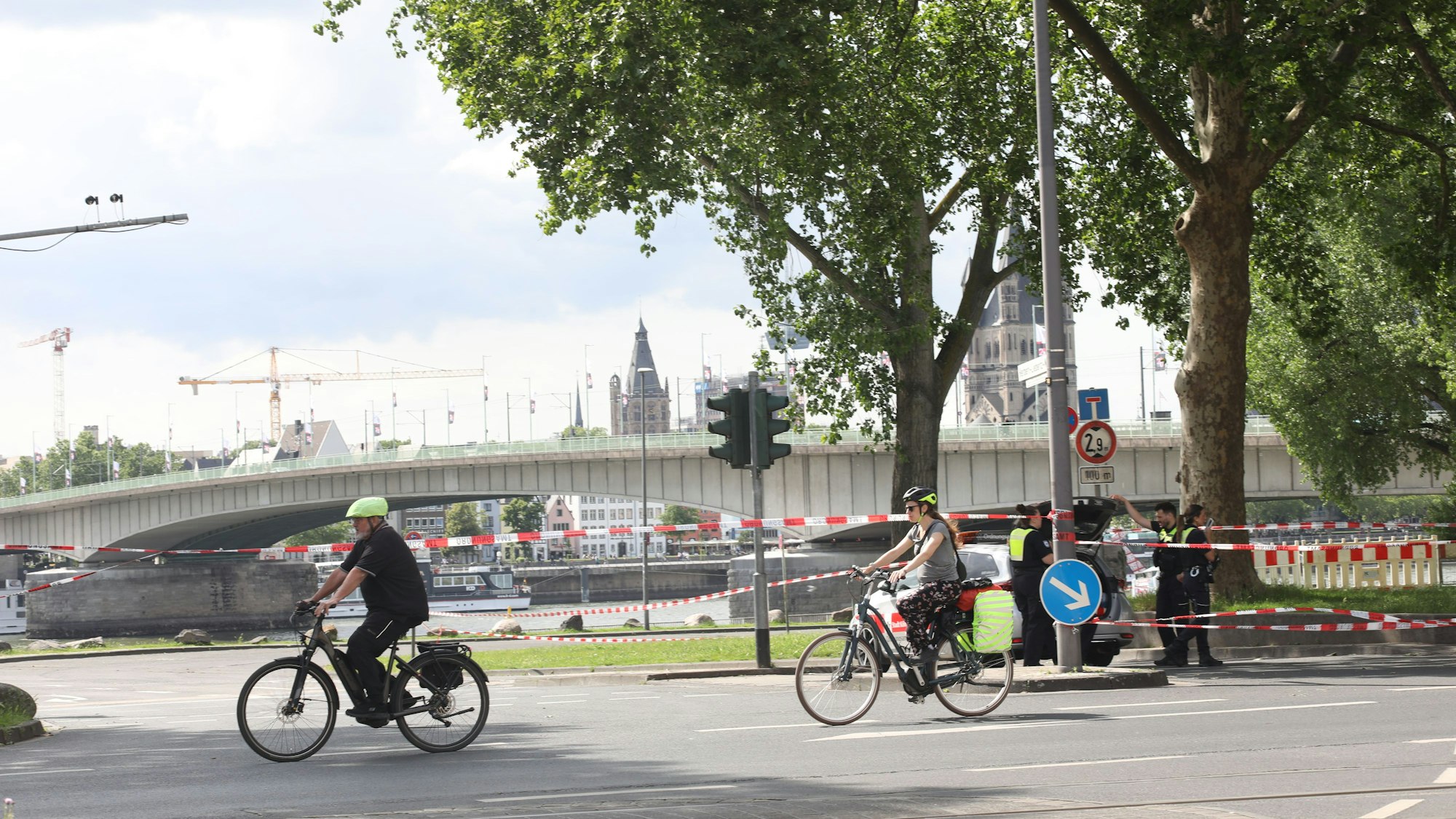 Fahrradfahrende radeln an einem abgesperrten Bereich in Rheinnähe vorbei. Im Hintergrund führt eine Brücke über den Fluss.